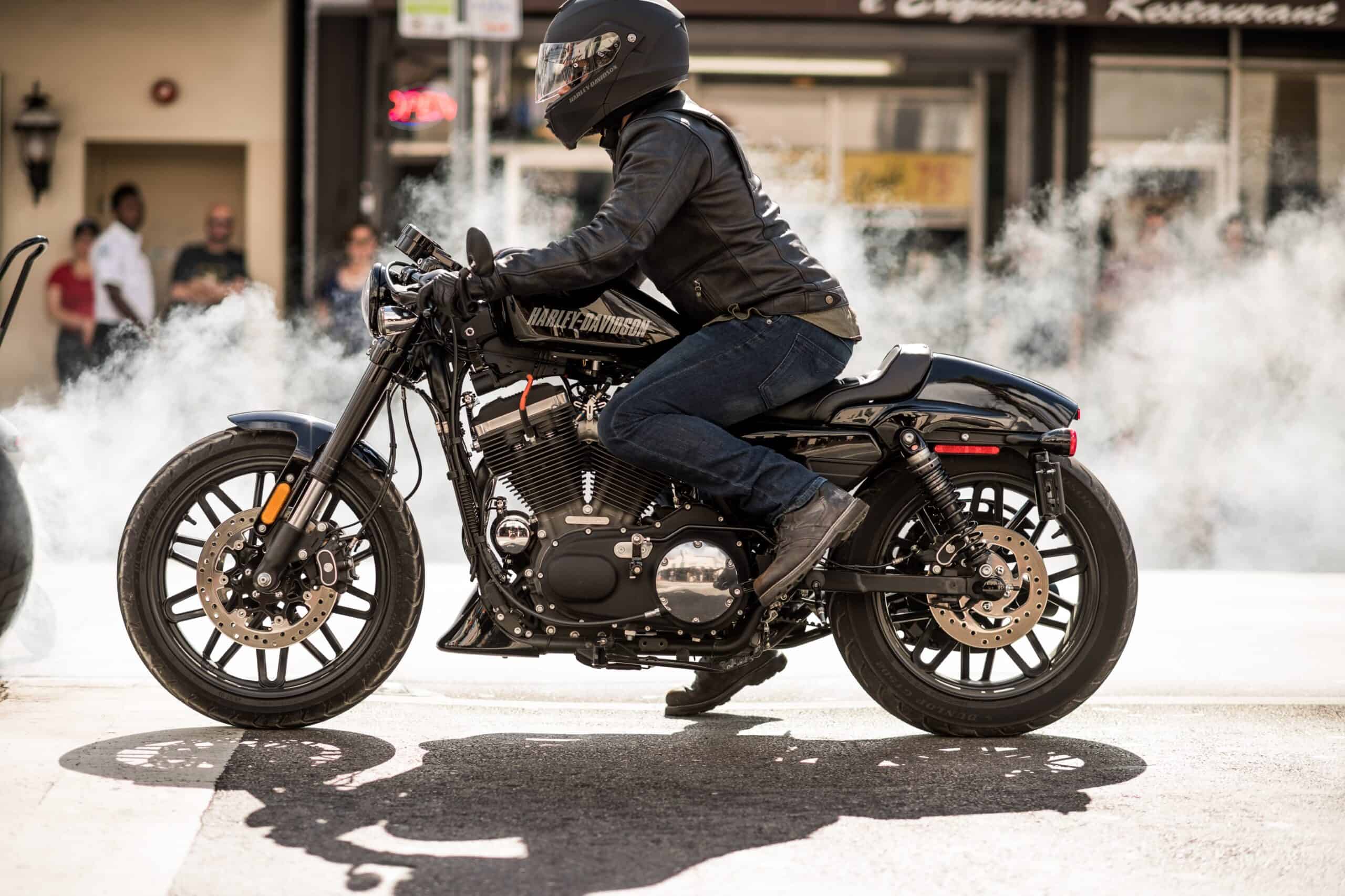 A motorcyclist in a black helmet and leather jacket rides a black motorcycle, with white smoke trailing behind