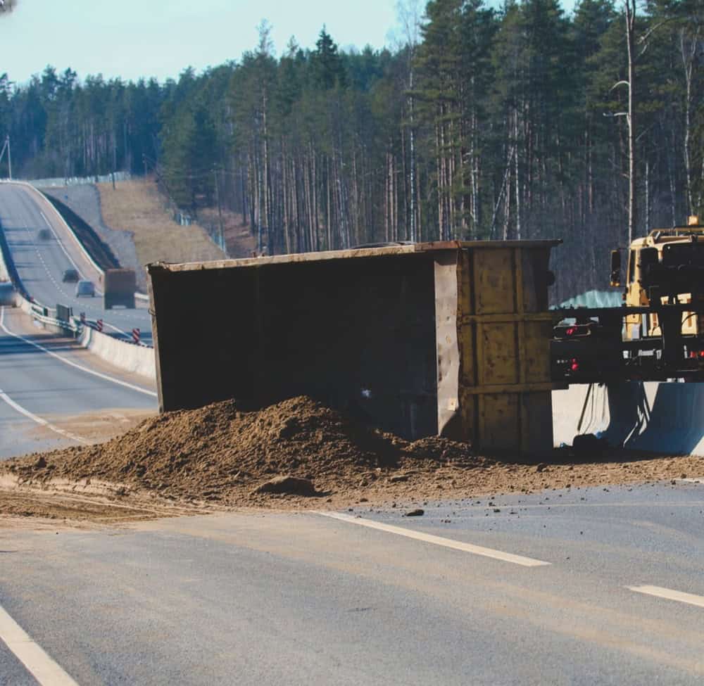 Dump truck accident on the highway with spilled dirt blocking the road.