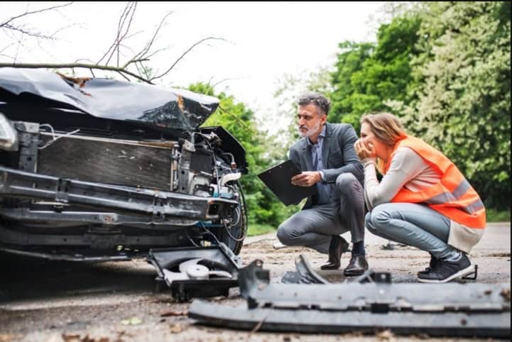 Man inspecting damaged car after crash while woman in safety vest looks concerned