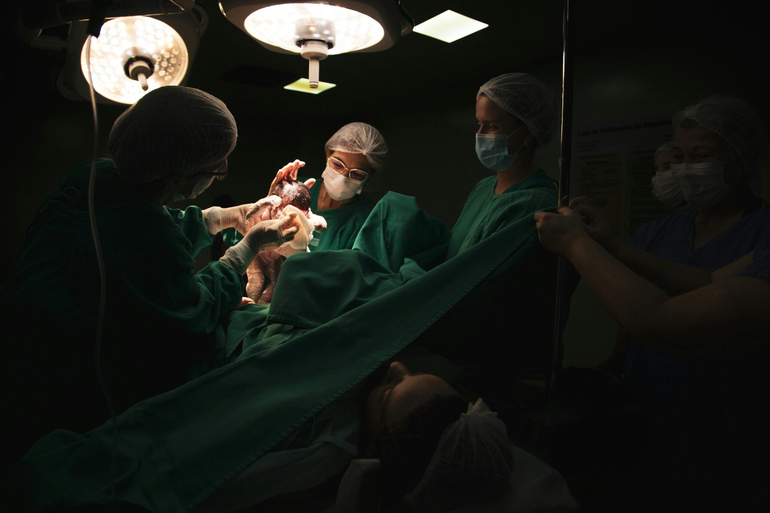 Medical team performing delivery in operating room, newborn emerging under surgical lights
