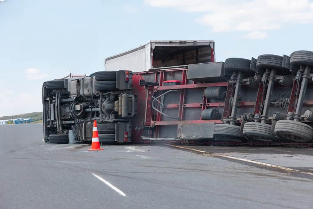 Overturned truck accident on highway with cargo trailer flipped and traffic cone nearby.