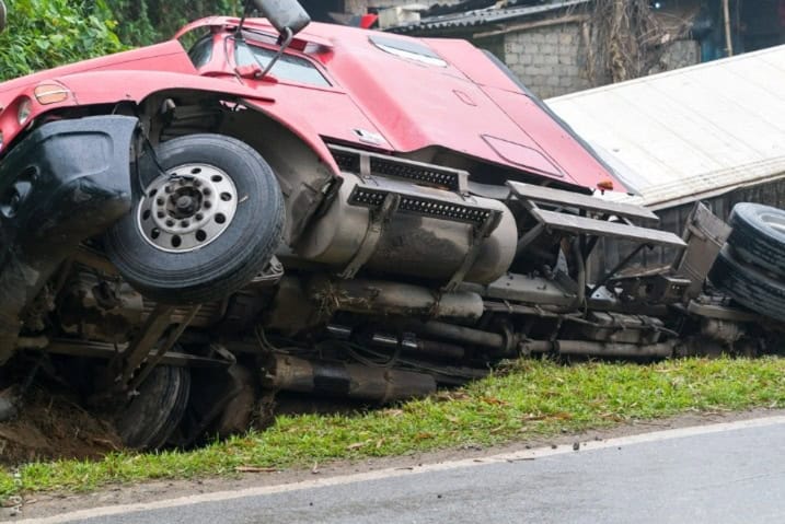Overturned red semi truck lies on roadside after rollover crash with visible damage.